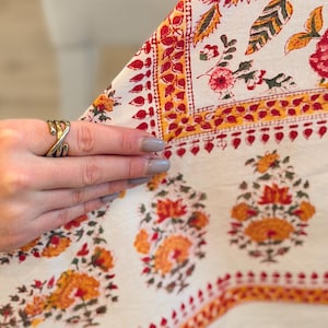 May include: Cream-colored textile with a floral pattern in red, orange, and yellow. The design includes stylized flowers and leaves, with a border featuring small red shapes. The fabric is folded, revealing the pattern's repeat.
