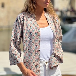 May include: A light brown, button-down shirt with a floral pattern in red, green, and purple. The shirt is worn over a white tank top and paired with white jeans. The sleeves are three-quarter length.