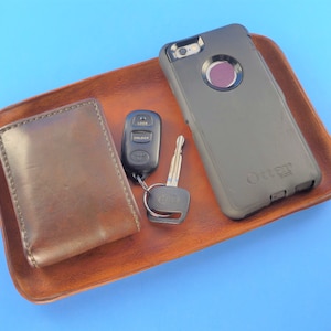 Pode incluir: Uma bandeja de valet de couro marrom com uma carteira de couro marrom, uma capa de telefone preta com o logotipo OtterBox, uma chave de carro com um controle remoto preto e uma chave prateada.