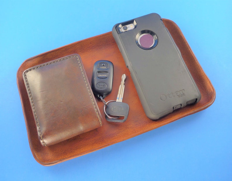 Pode incluir: Uma bandeja de valet de couro marrom com uma carteira de couro marrom, uma capa de telefone preta com o logotipo OtterBox, uma chave de carro com um controle remoto preto e uma chave prateada.
