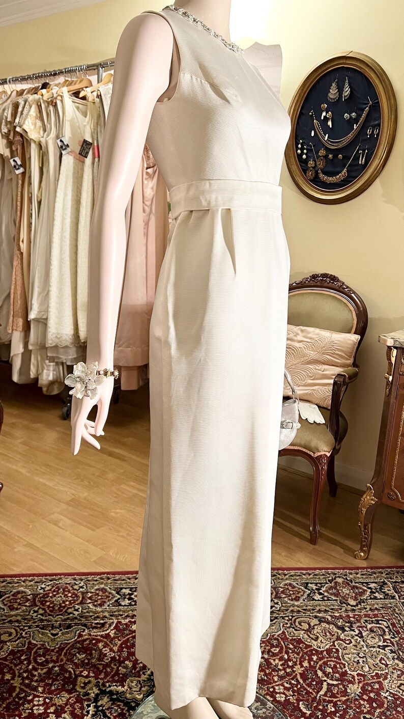 May include: A white sleeveless maxi dress with a beaded neckline and a fitted bodice. The dress has a simple, elegant design and is perfect for a formal occasion.
