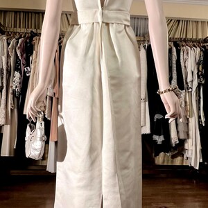 May include: A white sleeveless dress with a low back and a beaded neckline. The dress has a fitted bodice and a flowing skirt.