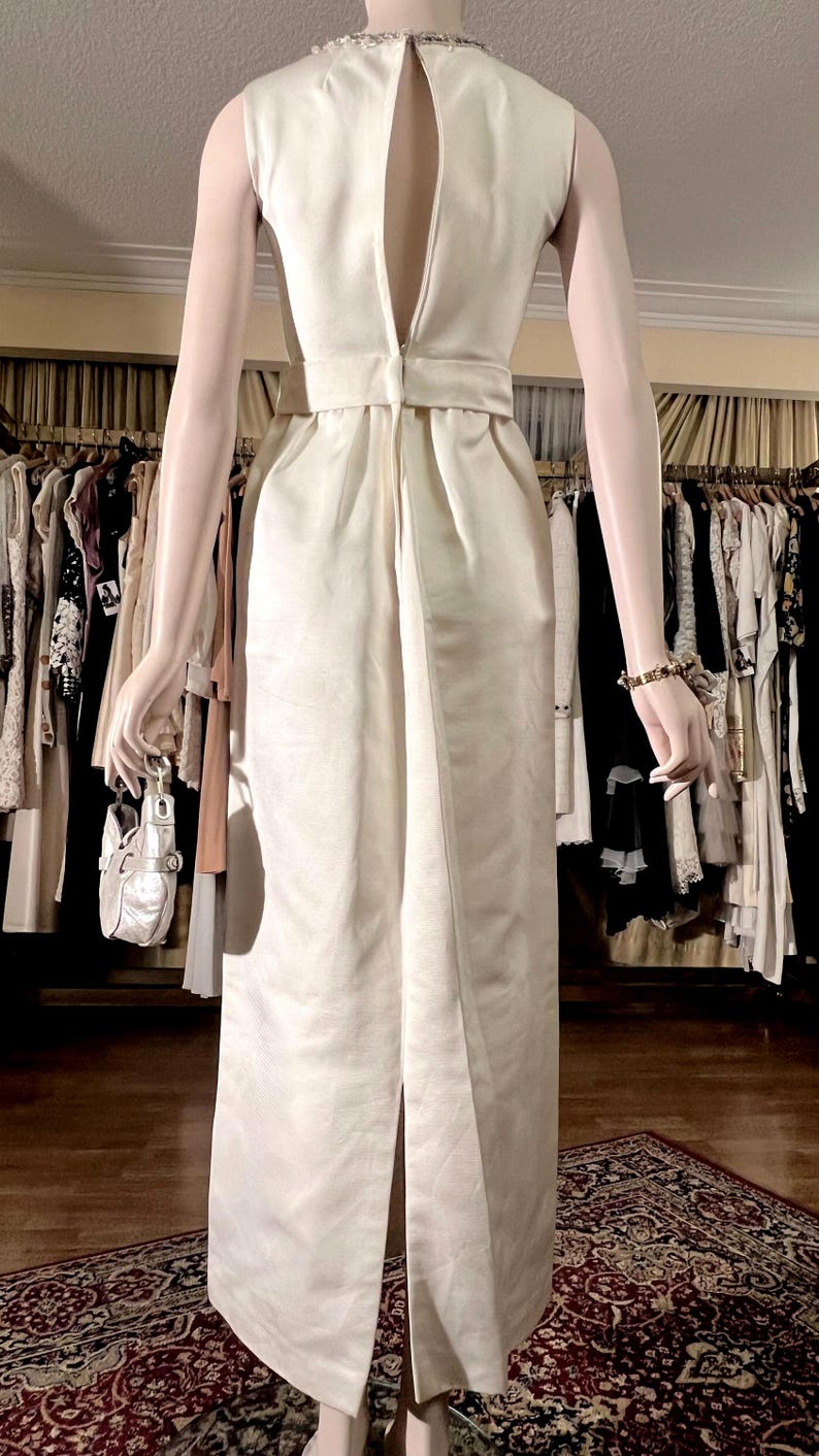 May include: A white sleeveless dress with a low back and a beaded neckline. The dress has a fitted bodice and a flowing skirt.