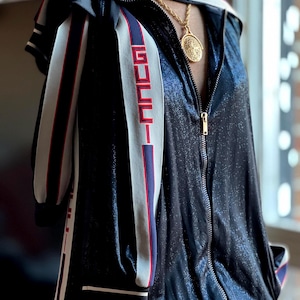 May include: A black zip-up jacket with white and red striped accents on the sleeves. The jacket has a white band across the shoulders with the word "GUCCI" in red. A gold chain with a pendant is visible.