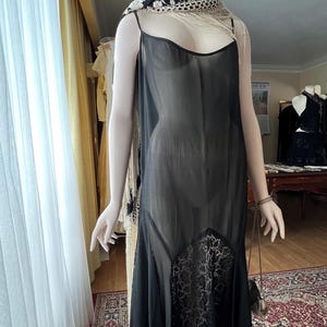 May include: A black vintage slip dress with spaghetti straps and a lace panel at the hem. The dress is displayed on a mannequin, paired with a black and white scarf and a feathered headpiece. The dress is sheer and has a flowing silhouette.