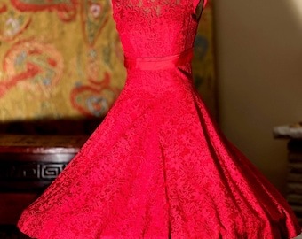 Puede incluir: Un vestido rojo de encaje con una cinta de satén. El vestido tiene una falda completa y un corpiño ajustado. El vestido está en un maniquí.