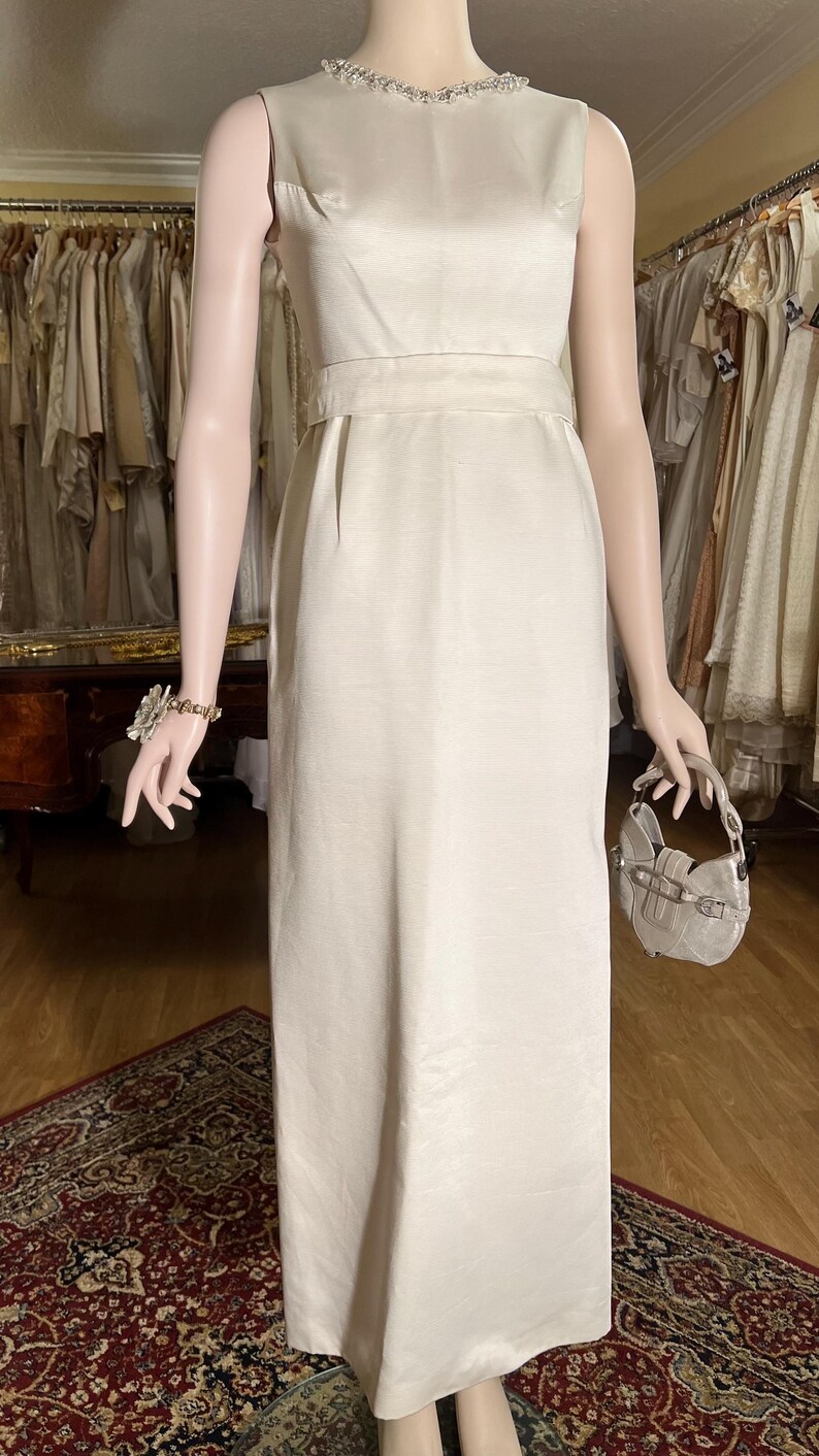 May include: A sleeveless white satin dress with a beaded neckline and a matching sash. The dress has a simple, elegant design and is perfect for a formal occasion.
