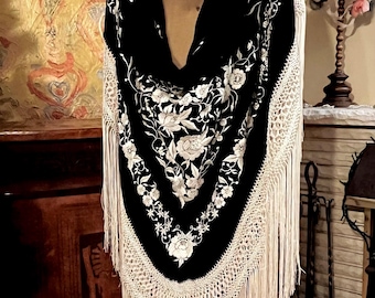 May include: A black shawl with white floral embroidery and a cream-colored fringe. The shawl is draped over a mannequin, showcasing the intricate design and texture. The floral pattern is detailed and covers the entire shawl.