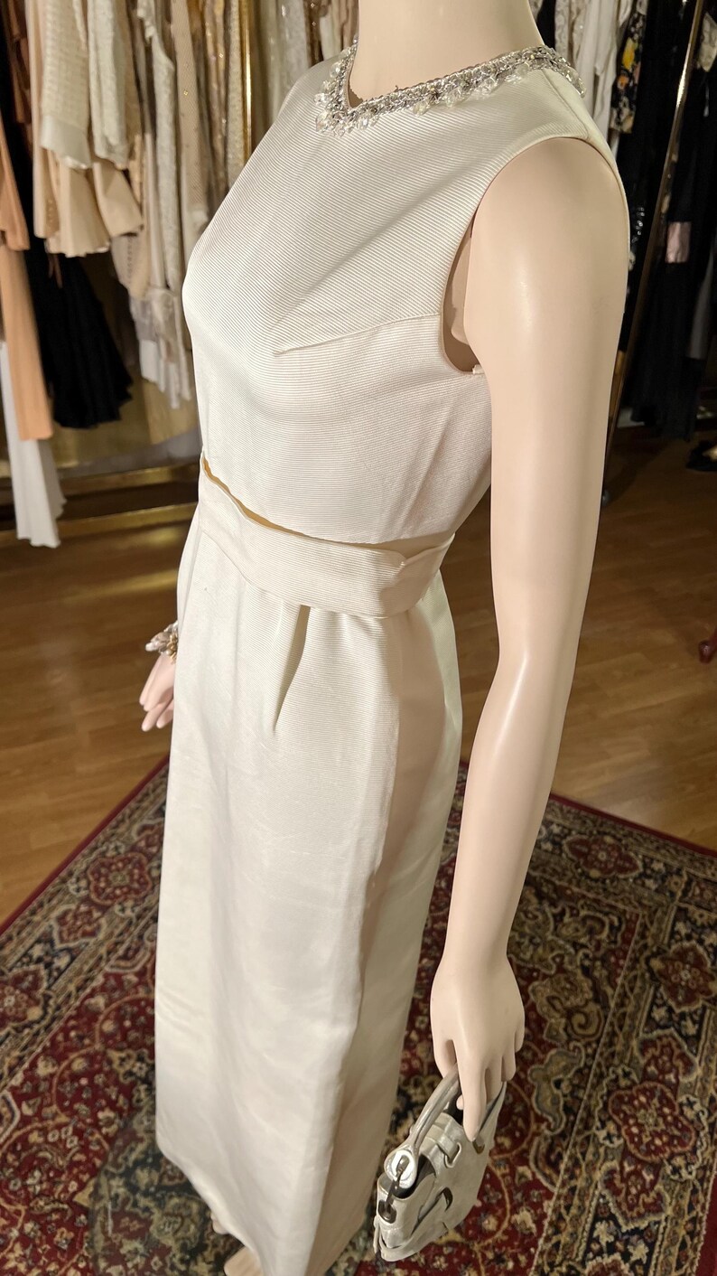 May include: A white sleeveless dress with a beaded neckline and a fitted silhouette. The dress has a belt at the waist and a straight skirt.