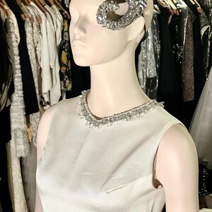 May include: A white sleeveless dress with a beaded neckline and a silver jeweled headpiece. The dress is on a mannequin in a clothing store.