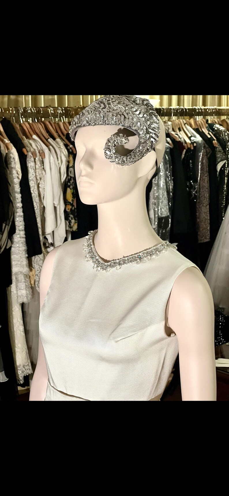 May include: A white sleeveless dress with a beaded neckline and a silver jeweled headpiece. The dress is on a mannequin in a clothing store.