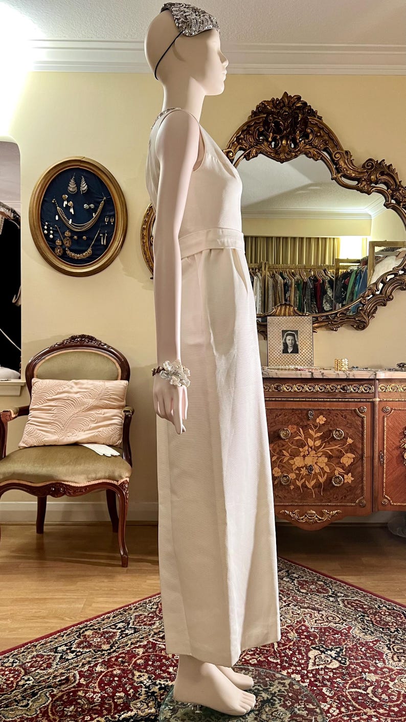 May include: A white sleeveless jumpsuit with a V-neckline and a wide leg. The jumpsuit is made of a lightweight fabric and has a relaxed fit. The model is wearing a silver headband.