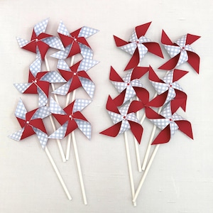 May include: A collection of eight pinwheels arranged in two groups. Each pinwheel features alternating blades of solid red cardstock and light blue and white gingham patterned paper, all mounted on thin white sticks with a small center fastener.