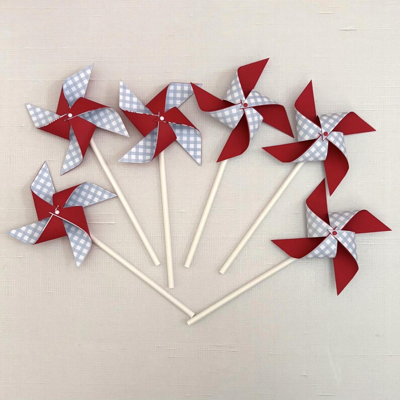 May include: Five pinwheels arranged in a fan shape, each featuring four alternating blades of solid red cardstock and white and light gray gingham patterned paper. Each pinwheel is attached to a thin, white, cylindrical paper stick.