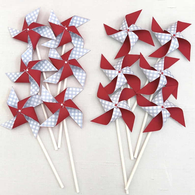 May include: A collection of eight pinwheels arranged in two groups, each featuring a four-pointed design with alternating solid red and light blue gingham patterned paper blades attached to thin white sticks with a small white center fastener.