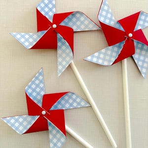 May include: Three red and blue pinwheels with white gingham patterns on the blades. The pinwheels are attached to white sticks.