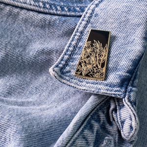 May include: A gold-colored enamel pin with a black background featuring a detailed floral design. The pin is attached to a denim jacket.