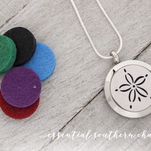 May include: A silver necklace with a round pendant featuring a cutout of a sand dollar. The necklace comes with five felt pads in green, black, blue, purple, and red.