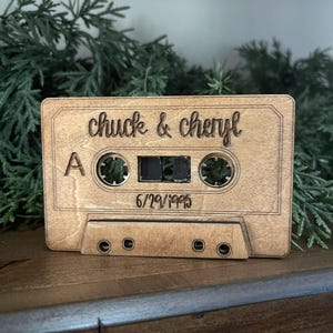 May include: A wooden cassette tape with the names "chuck & cheryl" and the date "6/29/1995" engraved on it. The tape is light brown and has the typical holes and details of a cassette. Green foliage is in the background.