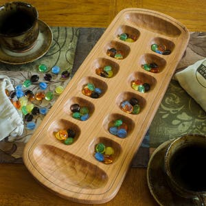 Mancala board made out of medium colored cherry wood, shown with multicolored glass playing pieces. Board is sanded smooth and small holes and larger stores show carving marks.