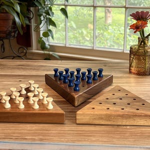 May include: Three wooden triangle-shaped board games with pegs. One board has white pegs, another has blue pegs, and the third is empty. The games are on a wooden table, with a vase of flowers in the background.
