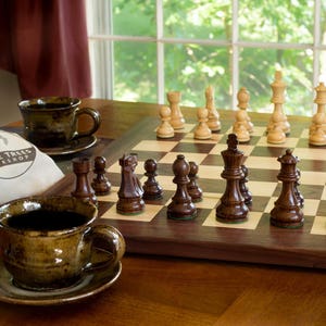 May include: A wooden chessboard with dark brown and light brown squares. The chess pieces are made of dark wood and are arranged on the board in a starting position. The chessboard is sitting on a wooden table with two brown ceramic coffee cups and saucers. A white canvas bag with the text "Three Trees Workshop" is in the foreground.