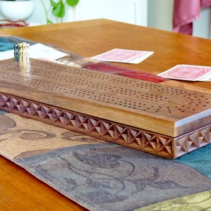 May include: A wooden cribbage board with a carved design on the edges. The board is closed and has a set of pegs in the top slot.