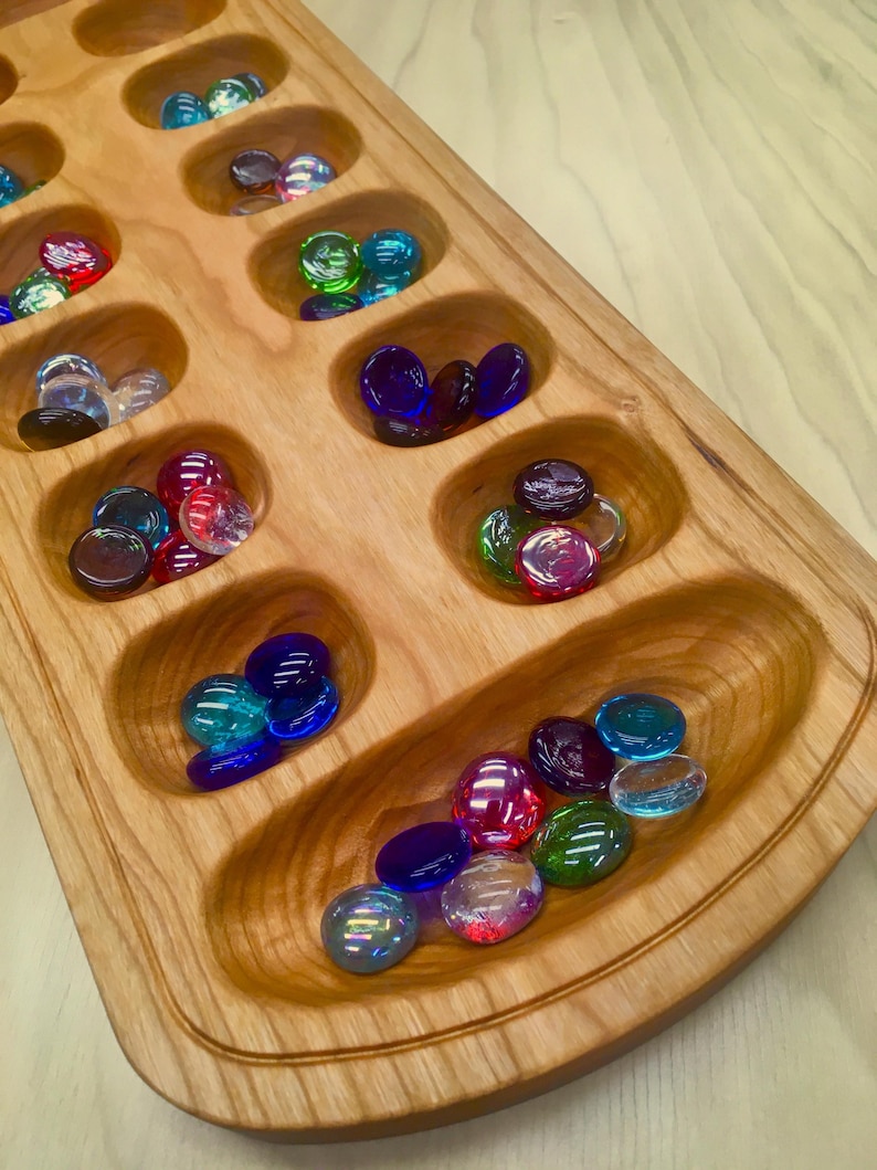 A wooden mancala board made from medium colored cherry wood. Each of the small pits and larger stores hold multicolored glass playing pieces.