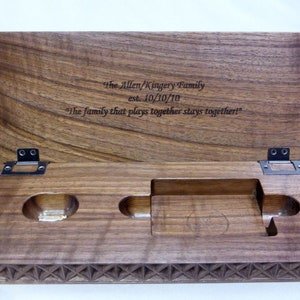 A dark walnut wood cribbage board with hinged lid is open. Thre is a custom engraved message centered on inside of the lid. There are spaces cut out of the base to hold cribbage pegs and a deck of cards.