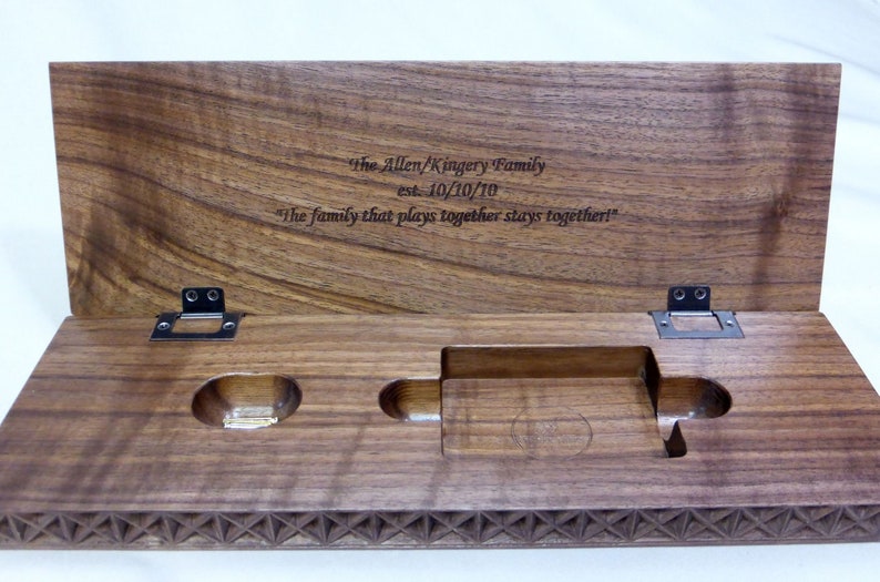 A dark walnut wood cribbage board with hinged lid is open. Thre is a custom engraved message centered on inside of the lid. There are spaces cut out of the base to hold cribbage pegs and a deck of cards.