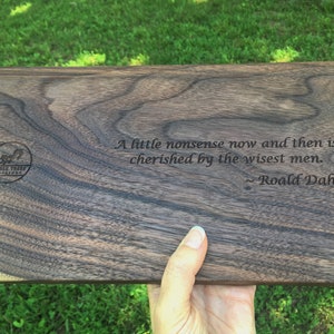 Custom engraved message on back of a walnut mancala board is a quote from Roald Dahl: A little nonsense now and then is cherished by the wisest men. The board itself is a dark brown with sweeping grain pattern.