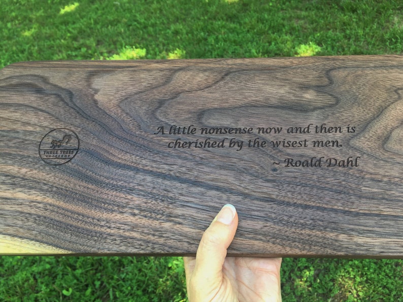 Custom engraved message on back of a walnut mancala board is a quote from Roald Dahl: A little nonsense now and then is cherished by the wisest men. The board itself is a dark brown with sweeping grain pattern.