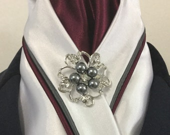 May include: A formal ascot in white satin with a burgundy center and a silver and pearl brooch. The ascot has a gray and burgundy trim. The brooch is a silver flower with gray pearls and a central clear gem.