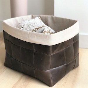 Puede incluir: Una cesta de almacenamiento de cuero marrón oscuro con un diseño tejido. La cesta tiene un forro interior de tela color crema y un borde doblado. Un artículo con estampado blanco y dorado está dentro de la cesta. La cesta está sobre un suelo de madera clara.