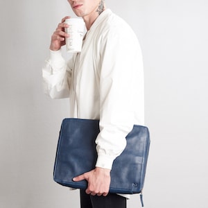 May include: A navy blue leather laptop case with a zipper closure. The case is rectangular and has a textured surface. The person in the image is holding the case and a white coffee cup.
