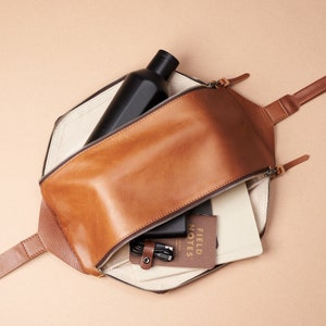 May include: A brown leather satchel bag with a zip closure. The bag is open and contains a black water bottle, a notebook, and a small pouch with cords. The notebook has the text "FIELD NOTES" printed on the cover.