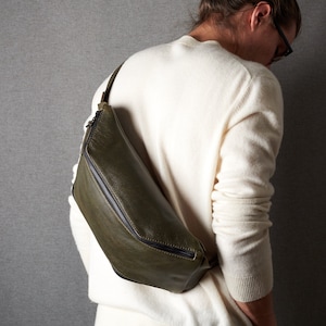 Peut inclure: Un sac à bandoulière en cuir vert foncé avec une fermeture éclair et une sangle noires. Le sac est porté par une personne portant un pull blanc.
