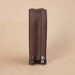 Puede incluir: Una bolsa de cuero marr&oacute;n con cremallera con dos cremalleras. La bolsa est&aacute; de pie sobre una superficie marr&oacute;n claro.