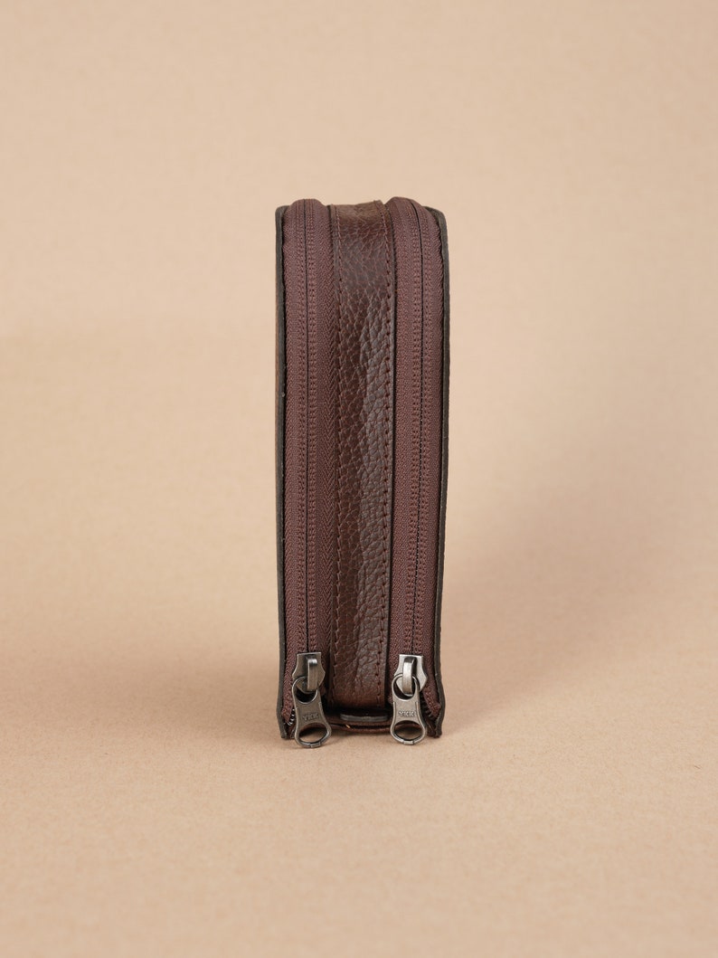 Puede incluir: Una bolsa de cuero marr&oacute;n con cremallera con dos cremalleras. La bolsa est&aacute; de pie sobre una superficie marr&oacute;n claro.