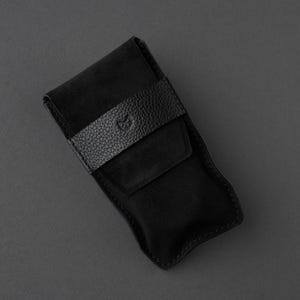 May include: A black leather case with a textured strap and a flap closure. The case is made of a soft, suede-like material and has a stitched edge. The strap features a small embossed design. The case is designed to hold small items.