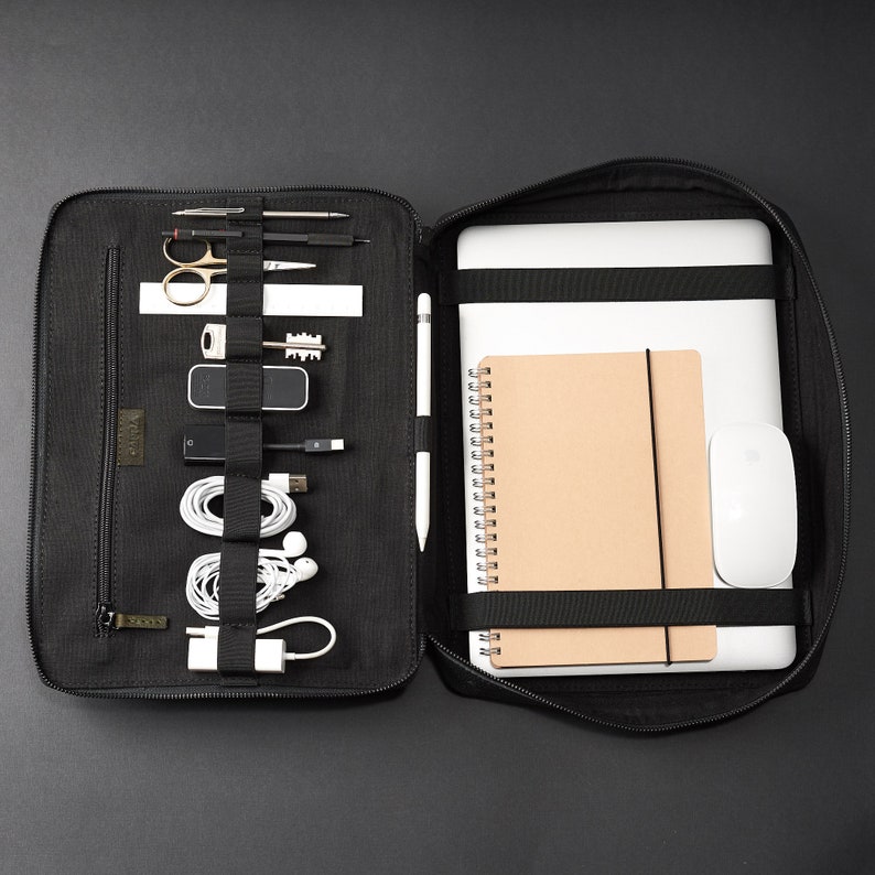 May include: A black canvas laptop bag with a zippered compartment and multiple pockets. The bag is open to reveal a laptop, a notebook, a mouse, and various other items, including pens, pencils, and cords.