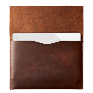 May include: A brown leather sleeve for a laptop or tablet. The sleeve has a flap that folds over the top and a small pocket on the front. The sleeve is made of a high-quality leather and has a distressed look.