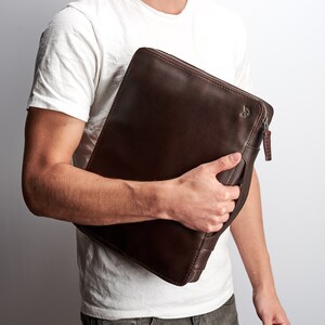 May include: A brown leather portfolio with a zipper closure and a small logo on the front. The portfolio is being held by a person.