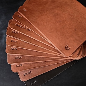 May include: A stack of brown leather coasters with names embossed on each one. The names include:  EFP, TCS, DANIELLE N LEE, JERZY, SHAY, JOHN, JACK, ALEX, BRIAN, and RCH.
