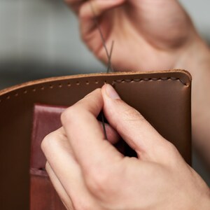 May include: Close-up of a person's hands stitching brown leather with a needle and thread. The leather is being sewn together to create a new item.