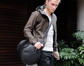 May include: A man wearing a brown jacket and black jeans is carrying a black leather duffel bag over his shoulder.