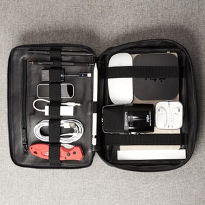 May include: Black leather carrying case with multiple compartments and elastic straps. The case is open and contains a variety of items, including a microphone, headphones, a knife, and a stylus.