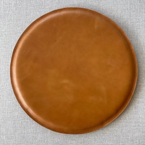 Könnte beinhalten: Ein runder, hellbrauner Leder-Tablett mit genähtem Rand. Das Tablett hat eine warme, mittelbraune Farbe und scheint aus hochwertigem Leder gefertigt zu sein. Die kreisförmige Form und das schlichte Design deuten auf Funktionalität und Stil hin.