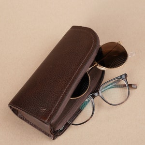 Puede incluir: Un estuche para gafas de cuero marr&oacute;n con cierre de cremallera. El estuche est&aacute; abierto y hay dos pares de gafas dentro. Un par son gafas de sol redondas con lentes marrones y el otro par son gafas rectangulares con lentes transparentes.