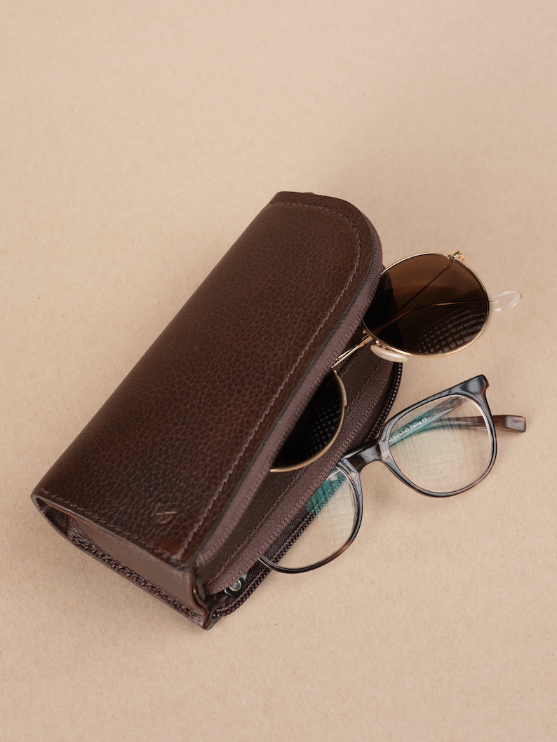 Puede incluir: Un estuche para gafas de cuero marr&oacute;n con cierre de cremallera. El estuche est&aacute; abierto y hay dos pares de gafas dentro. Un par son gafas de sol redondas con lentes marrones y el otro par son gafas rectangulares con lentes transparentes.